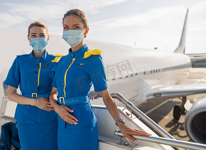 两位身穿蓝色制服、戴着防护面罩的漂亮空中小姐看着镜头，在登机时站在楼梯上
