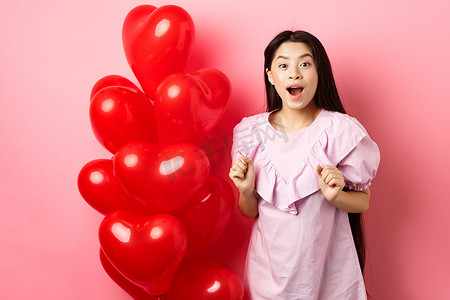 很奇怪摄影照片_兴奋而惊讶的韩国少女张开嘴巴，在情人节那天收到惊喜礼物，看起来很奇怪，站在心形气球旁边，粉红色背景