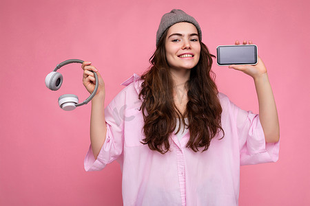 照片中，美丽正面微笑的年轻女性身穿时尚休闲装，与彩色背景墙隔开，手持白色蓝牙无线耳机，展示手机屏幕为空，用于模拟看相机