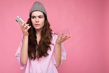 情绪集中、迷人的黑发年轻女性穿着时尚的粉色衬衫和灰色帽子，与粉色背景隔离，手牵手，使用手机交流和收听语音信息