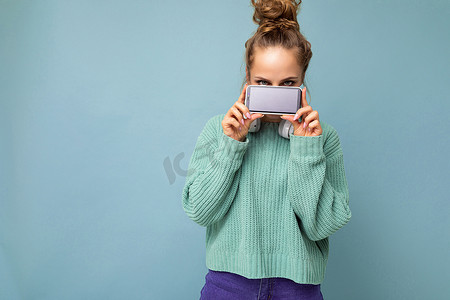 手指拿着手机摄影照片_照片中，漂亮的年轻女性穿着休闲装，站在背景中，与世隔绝，复印空间拿着智能手机，手里拿着手机，屏幕显示为空，用于模拟
