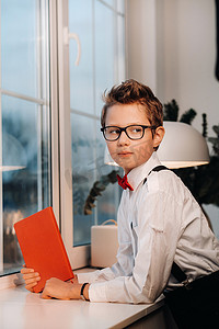 窗边站着一个男孩，他身穿衬衫，系着红色领结，戴着眼镜，手里拿着一本红色的书