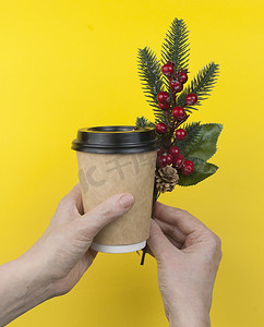 手拿着一次性咖啡杯，黄色背景上有圣诞浆果树枝。
