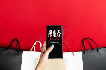 红色创意促销摄影照片_黑色星期五促销，女性在智能手机上进行网上购物，红色背景上有购物袋
