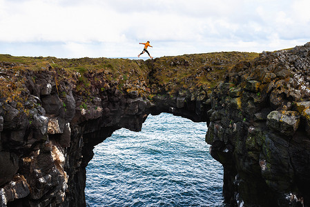 阿尔纳斯塔皮摄影照片_游客跳过冰岛阿尔纳斯塔皮的一座天然石桥
