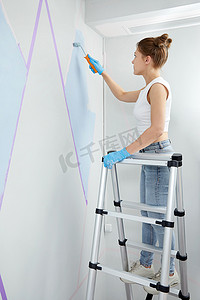 年轻女子用油漆滚筒刷墙，站在梯子上用遮蔽胶带