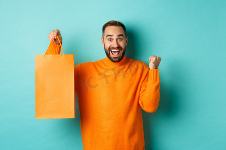 快乐的男人拿着橙色购物袋欢欣鼓舞，打折庆祝，站在绿松石色的背景下