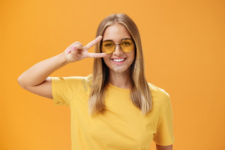 金黄标志摄影照片_可爱乐观友善的年轻白人女性，头发金黄，身穿黄色 T 恤，戴着太阳镜，近眼展示和平标志，在橙色背景下对着镜头欢快地微笑