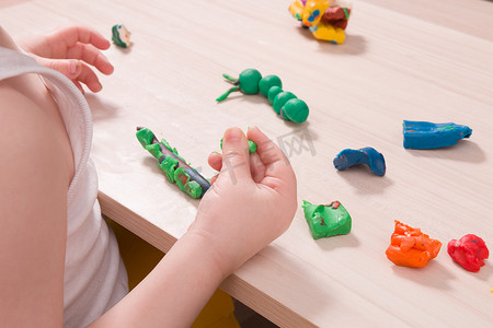 动漫雕刻摄影照片_一个小孩在木桌上用绿色橡皮泥雕刻毛毛虫，手部精细运动技能的发展，在家里和孩子玩耍，前景中用橡皮泥雕刻毛毛虫