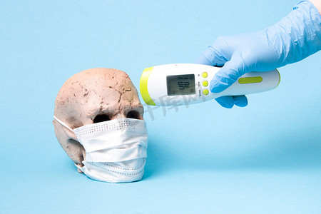 陶瓷头骨面具和手戴一次性医用橡胶蓝色手套，蓝色背景红外非接触式温度计，复印空间，高温40摄氏度