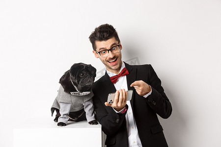 快乐的狗主人在手机上展示一些东西给宠物，男人和哈巴狗穿着华丽的服装，站在白色背景上