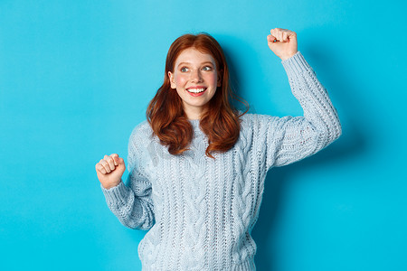 快乐的红发女孩为团队加油，举手欢呼，微笑，庆祝胜利或成功，站在蓝色背景下