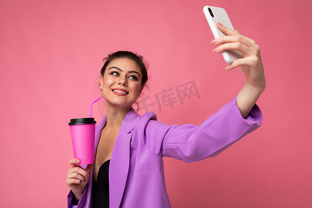 在工作室的粉红色背景中，一位身着时尚紫色西装、微笑着的年轻黑发美女的特写，她拿着一部流行的手机，拍着自拍照片，喝着吸管饮料