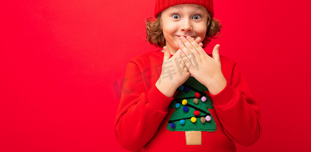 穿着红色圣诞毛衣的酷少年在红墙、温暖的帽子和带圣诞树的毛衣的背景下闲逛