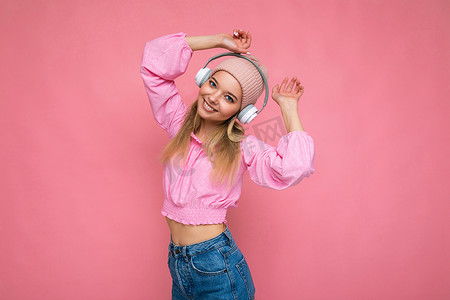 照片中，身穿粉红色衬衫和粉红色帽子的迷人正面微笑的年轻金发女性在粉红色背景墙上隔开，戴着白色无线蓝牙耳机听音乐玩得开心，看着相机