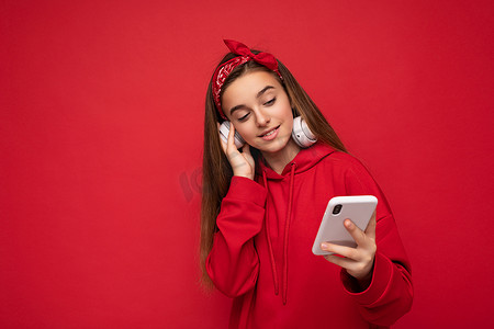 照片中，身穿红色连帽衫的美丽正面黑发女孩在红色背景中被隔离，手持并使用智能手机，戴着白色无线耳机，听着酷炫的音乐，看着 gadjet 显示器