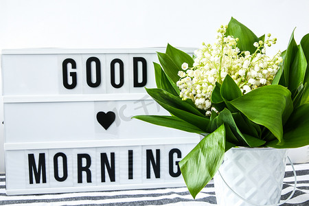 文字早上好摄影照片_带有文字“早上好”的灯箱和白色桶中的铃兰花束，春天
