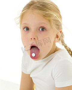 干净的女孩摄影照片_一个穿着干净的白色 T 恤的小女孩吞下一颗药丸，药丸在她的舌头上。 