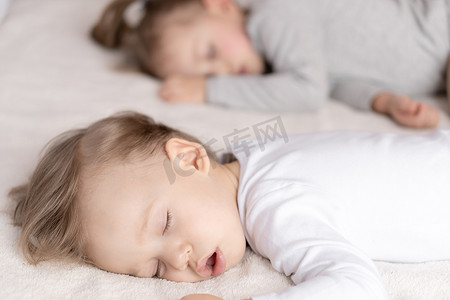 孩子和妈妈睡摄影照片_童年、睡眠、放松、家庭、生活方式概念 — 两个 2 岁和 3 岁的幼儿穿着白色和米色紧身衣，午餐时睡在米色和白色床上，手牵手俯视。