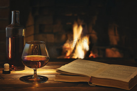 燃烧的壁炉旁的桌子上放着一杯白兰地、一瓶酒和一本打开的旧书。