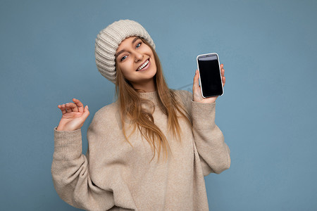 美丽正面漂亮的年轻女性穿着时尚的米色毛衣和米色针织冬帽的照片，在蓝色背景中与世隔绝，手里拿着空的空间，并展示带有空屏幕的手机，用于模拟看相机