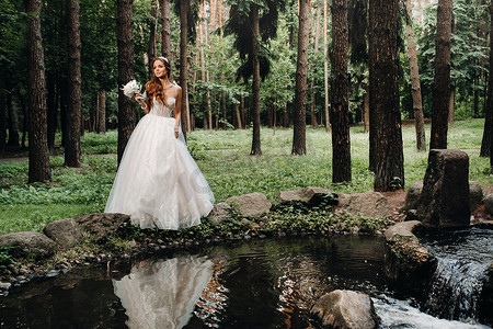 小溪边摄影照片_一位身着白色连衣裙、戴着手套、手持花束的优雅新娘站在森林里的小溪边，享受着大自然。自然公园里穿着婚纱和手套的模特。白俄罗斯