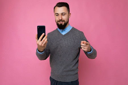 留着胡子、穿着时髦的灰色毛衣和蓝色衬衫的英俊、快乐、酷酷、胡子拉碴的年轻黑发男人站在粉红色的背景墙上，拿着智能手机，用手机看着 gadjet 指着你