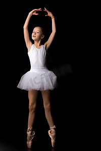 可爱芭蕾摄影照片_穿着芭蕾舞短裙和足尖鞋的可爱小女孩正在演播室里以黑色背景跳舞。