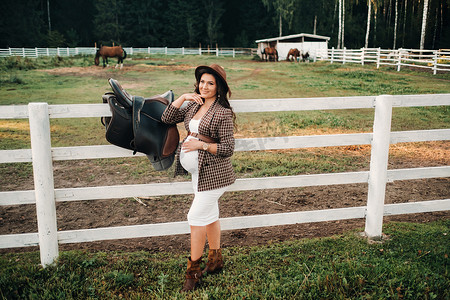 一个怀孕的女孩，在大自然中的一个围场附近的马旁边戴着一顶帽子，肚子很大。穿着棕色连衣裙的时尚孕妇与马。