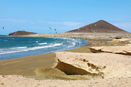 许多五颜六色的风筝在海滩上，风筝冲浪者在大风天在特内里费岛的加那利群岛 El Medano 乘风飞翔，地平线上有蒙大拿罗亚山