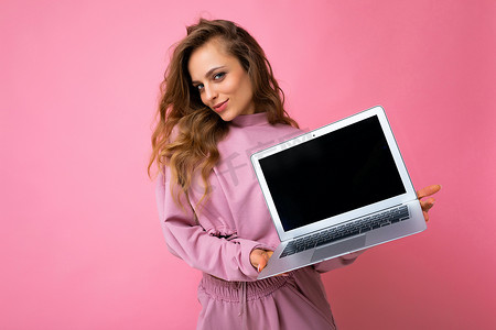 粉色墙壁摄影照片_照片中，美丽的金发卷发年轻女子看着相机，手里拿着电脑笔记本电脑，笔记本电脑上有空的显示器屏幕，上面有模拟和复制空间，穿着粉色连帽衫，与粉色墙壁背景隔离