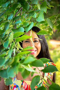 一位美丽的年轻混血非洲裔美国女性穿着黄色花卉印花连衣裙，从树枝后面露出牙齿微笑，树叶遮住了她的脸和上半身。