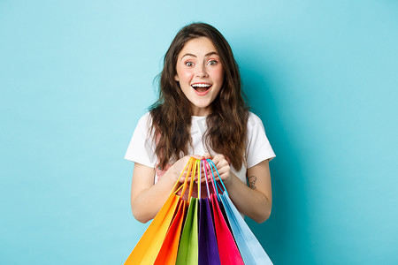 兴奋的女购物者的画像，拿着购物袋，在商店购物，惊讶地微笑，享受折扣，站在蓝色背景上