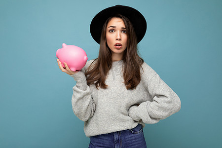 震惊惊讶的年轻美丽女人的画像，深色头发穿着时尚的灰色毛衣和黑色帽子隔离在蓝色背景与空空间和拿着粉红色的小猪盒钱。