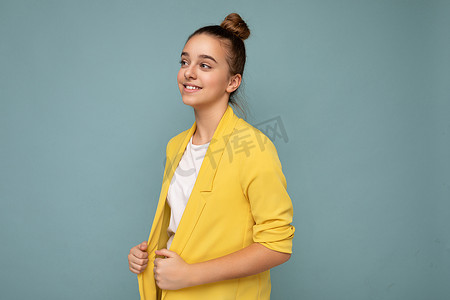 侧面照片中，迷人、积极、快乐、微笑的黑发小女孩穿着休闲黄色夹克和白色 T 恤，站在蓝色背景墙上，看着旁边