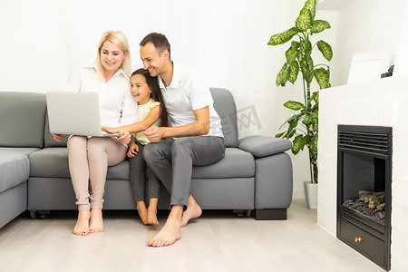 快乐的家庭，有小女孩一起坐在沙发上玩笔记本电脑，父母和小女儿在家里笑着放松，用电脑看有趣的互联网视频，打在线电话