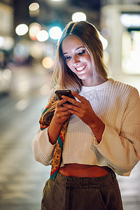 晚上看手机摄影照片_城市灯光散焦的女人看着她的智能手机。