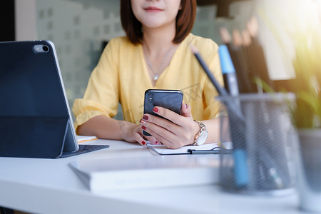 年轻的亚洲女商人在办公室的木桌上使用智能手机联系客户。