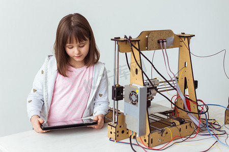 学校建模摄影照片_戴着 3d 打印百叶窗的可爱女孩正在看着她的 3d 打印机打印她的 3d 模型。