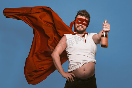 高能英雄摄影照片_喝醉的超级英雄或反英雄拿着一瓶酒，戴着超级英雄红色面具、戴着飞行斗篷的男人拿着一瓶酒，同时露出大肚子，在褪色的牛仔蓝色背景中被隔离