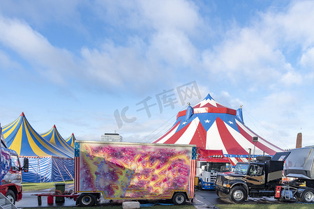 马戏团大篷车在红白相间的马戏团帐篷前，顶上是蓝星覆盖，映衬着阳光明媚的蓝天和云彩