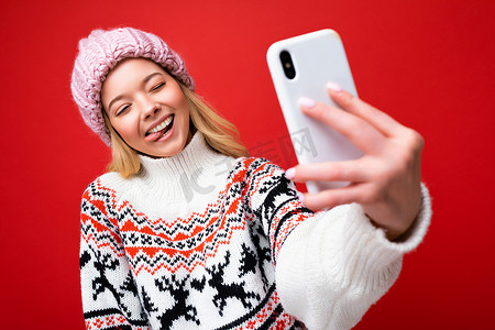 一张漂亮正面的年轻金发女性戴着温暖的针织帽和冬季温暖的毛衣站在红色背景中孤立地使用手机自拍照片看着设备显示屏并展示舌头的照片