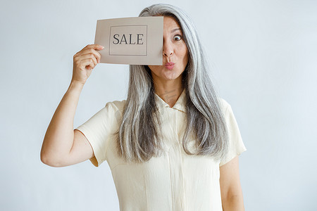 头发灰白的风趣成熟女人用浅灰色背景上的销售标志遮住了眼睛