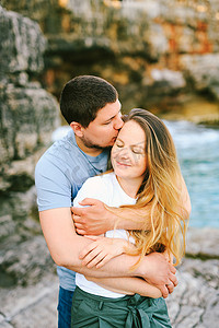 丈夫从后面拥抱并且在岩石岸和水的背景下亲吻微笑的妻子