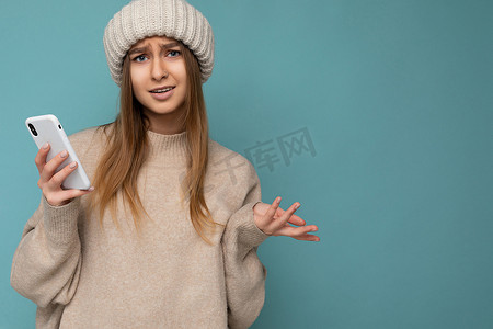 美丽不满的年轻金发女子的特写照片，她身穿时尚的米色保暖毛衣，头戴蓝色背景中突显的冬季保暖米色针织帽，手拿空地，使用手机短信看着相机和