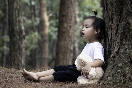 夹娃娃机边框摄影照片_亚洲小女孩抱着熊，独自坐在松树下的地上。