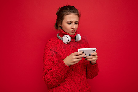 穿着红色毛衣的迷人、心烦意乱的深色年轻女性被隔离在红色背景墙上，手持和使用手机在互联网上冲浪，戴着蓝牙耳机看着 gadjet