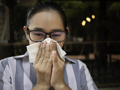 亚洲年轻女子拿着纸巾擦着眼镜，擤鼻涕。