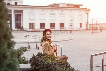 穿着秋装和米色的漂亮法式女人坐在大理石花坛上，在背景上分享或租用电动滑板车，等待她拿着咖啡杯的约会对象。