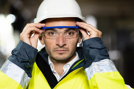 专业自信的严肃工程师看着相机、穿着安全制服和护目镜站在重工业工厂的特写镜头，准备在员工进行金属焊接时进行制造工作